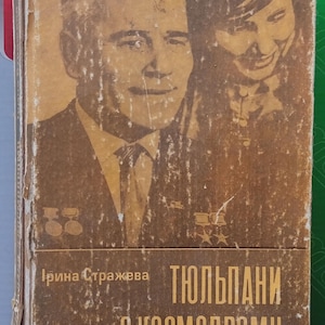 May include: A vintage hardcover book with a worn brown cover featuring a sepia-toned portrait of a man and a woman. The text on the cover reads "Ірина Стражева" and "ТЮЛЬПАНИ З КОСМОДРОМУ" in a light yellow font. The edges of the book show signs of wear.