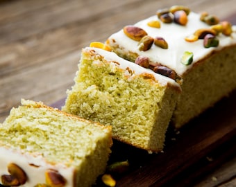 Puede incluir: Un bizcocho de pistacho con glaseado blanco y pistachos en la parte superior, cortado en tres trozos sobre una tabla de cortar de madera.
