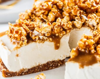 Puede incluir: Una porción de tarta de queso con salsa de caramelo y una cobertura de palomitas de maíz con caramelo. La tarta de queso tiene un relleno de color claro y una base de color marrón oscuro. Las palomitas de maíz con caramelo son una mezcla de colores dorados y marrones.