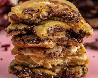 Puede incluir: Una pila de galletas recién horneadas con un exterior dorado y un rico relleno de chocolate. Las galletas están rellenas de chocolate derretido y malvavisco blanco, sobre un fondo rosa.