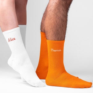 Male and female wearing vibrant embroidered unisex socks. Orange sock has white embroidery with male name and white sock showing female ankle with red embroidery