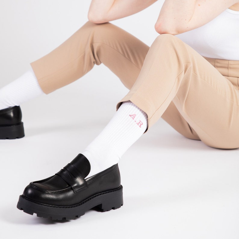 Woman wearing white ribbed cotton socks with pink initials embroidered on them. Style with black classic shoes and khaki trousers