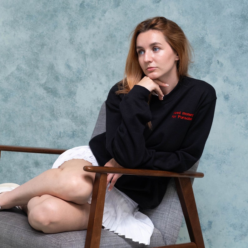 Young girl looks cute and cosy wearing black sweatshirt with personalised embroidery saying Need Money for Porsche. The sweatshirt is black and thread text is red