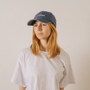 A young woman modelling a white t-shirt and vintage style unisex 6 panel cap in denim colour with custom embroidered text at the front saying happy thoughts