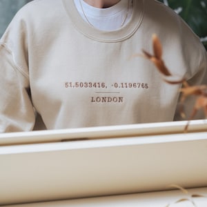 May include: A beige sweatshirt with the text "51.5033416, -0.1196765 LONDON" embroidered on the front.