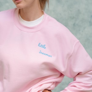 Könnte beinhalten: Ein hellrosa Sweatshirt mit den Worten "little dreamer" in hellblau gestickt. Das Sweatshirt hat einen Rundhalsausschnitt und wird über einem weißen T-Shirt getragen.