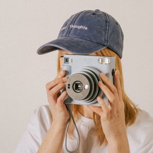 A boho style photo of a female wearing a custom made vintage style cap with personalised embroidery while taking a photo on Polaroid camera