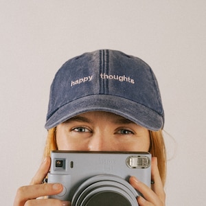A girl holding a Polaroid camera smiling and wearing a washed out vintage style denim cap with text saying happy thoughts, embroidered on the front