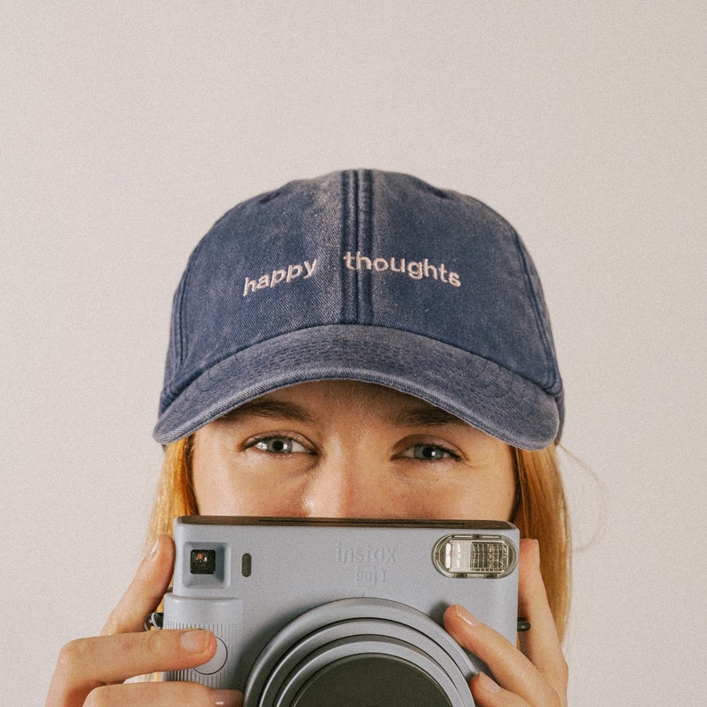 A girl holding a Polaroid camera smiling and wearing a washed out vintage style denim cap with text saying happy thoughts, embroidered on the front