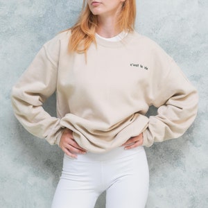 Female wearing light coloured unisex oversized sweatshirt with custom text embroidered on left chest of her pullover