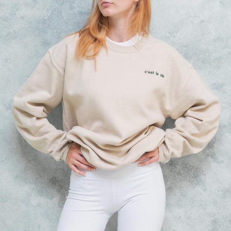 Female wearing light coloured unisex oversized sweatshirt with custom text embroidered on left chest of her pullover