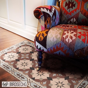 Può includere: Un tappeto a motivi geometrici in tonalità marrone, beige e blu. Il tappeto è posizionato su un pavimento in legno e una poltrona colorata con un tessuto a motivi è seduta sopra il tappeto.