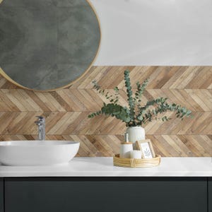 Op de afbeelding: Een witte keramische wastafel met een chromen kraan staat op een wit aanrecht. Achter de wastafel is een muur met een houtlook visgraat tegelpatroon. Een houten dienblad met een vaas met groene eucalyptus takken, een witte fles met een pomp en een witte kaars staat op het aanrecht.