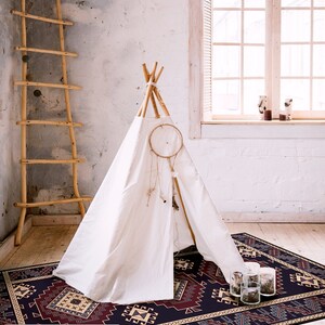 May include: A white teepee tent with a dreamcatcher hanging from the top, set on a dark blue and red patterned rug with beige accents. The rug has a geometric design and is in a room with a white brick wall and a wooden ladder.