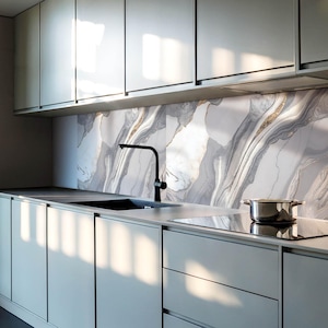 Op de afbeelding: Een moderne keuken met grijze kasten en een marmeren achterwand. De achterwand heeft een draaiend patroon in grijs, wit en goud. Een zwarte kraan is gemonteerd boven een roestvrijstalen gootsteen. Een zwart kooktoestel bevindt zich rechts van de gootsteen.