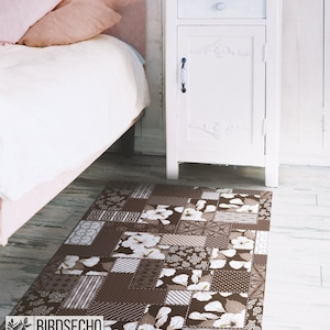 May include: A brown and white patterned area rug with a patchwork design featuring floral motifs. The rug is placed on a wooden floor in a bedroom setting.