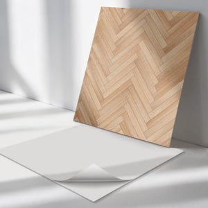 May include: A rectangular wooden panel with a herringbone pattern, leaning against a white wall. The panel is made of light brown wood. A white sheet of paper with a curled corner is in the foreground.
