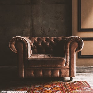 Op de afbeelding: Een bruine fluwelen Chesterfield-fauteuil met knoopdetails. De stoel staat op een houten vloer met een patroonkleed. De achtergrond is een donkerbruine muur. Het logo "BIRDSECHO" is zichtbaar op het kleed.