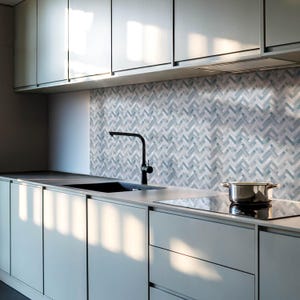 Puede incluir: Una cocina moderna con armarios grises y un salpicadero de azulejos en espiga gris y blanco. Un grifo negro está montado sobre un fregadero de acero inoxidable. Una placa de cocción eléctrica negra se encuentra a la derecha del fregadero.