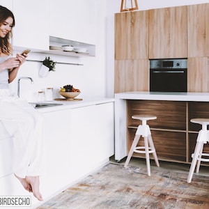 May include: A woman sits on a white kitchen counter, looking at her phone. The kitchen has white cabinets, a stainless steel sink, and a wooden cabinet with a built-in oven. The floor is covered in a patterned rug.