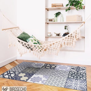Puede incluir: Una hamaca blanca con flecos cuelga en una habitación con paredes blancas y una estantería de madera. La hamaca tiene dos almohadas, una verde y otra con un diseño de hojas. Una alfombra azul y blanca con un diseño geométrico está en el suelo.