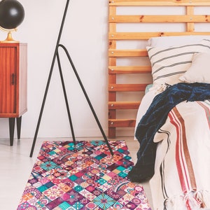 May include: A colorful patterned rug with a geometric design in shades of pink, purple, blue, and yellow. The rug is placed in a bedroom setting with a wooden bed frame and a black floor lamp.