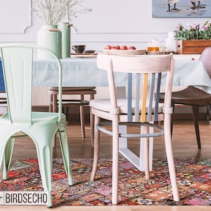 May include: A dining room with a light blue tablecloth, two light green metal chairs, a light wood chair with a gray and yellow striped back, and a brown leather chair. The chairs are arranged around a table with a colorful patterned rug underneath. The rug has a variety of colors and patterns, including red, orange, yellow, green, blue, and purple. The rug is made of a patchwork of different fabrics.