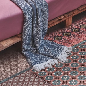 Può includere: Una coperta blu e bianca con motivo a frange sui bordi, drappeggiata su un letto rosa con una cornice in legno. Il letto è appoggiato su un tappeto con motivi multicolori e disegni geometrici.