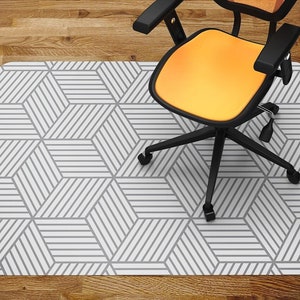 May include: A geometric patterned office chair mat in gray and white, with a black and orange office chair on top. The mat features a repeating pattern of angled lines, creating a 3D cube effect. The chair has a black base and an orange seat and back.