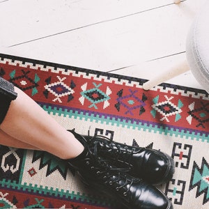 May include: A patterned rug with red, blue, and white geometric designs. A person is wearing black boots and sitting on the rug.