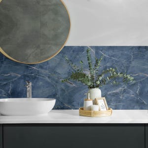 May include: A bathroom vanity with a white porcelain sink, a round mirror with a wooden frame, and a blue marble tile backsplash. A wicker tray holds a white vase with green eucalyptus branches, a white soap dispenser, and a white candle.