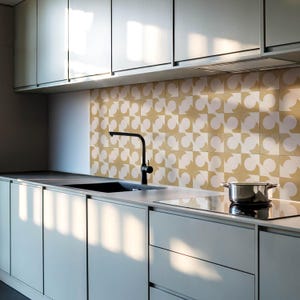 May include: A modern kitchen with white cabinets and a black faucet. The backsplash is made of white and yellow patterned tiles. A black stovetop and a stainless steel pot are on the counter.