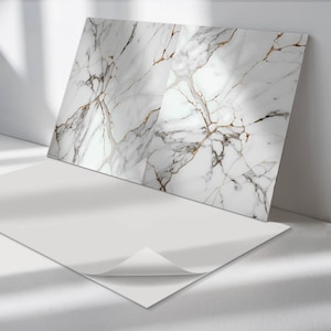 Puede incluir: Dos azulejos de mármol blanco con un patrón de vetas doradas. Los azulejos están sobre una superficie blanca.