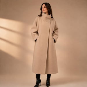 May include: A long, beige wool-blend coat featuring a high funnel neck, wide sleeves, and a straight, floor-length silhouette. The coat is worn over black trousers and black ankle boots, creating a clean, structured look with a smooth finish.