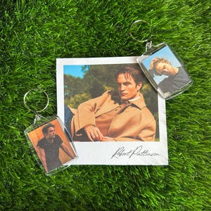 Könnte beinhalten: Zwei durchsichtige Acryl-Schlüsselanhänger mit rechteckigen Einlegern, die Fotos von Robert Pattinson zeigen, sowie ein quadratischer weißer Druck des Schauspielers in einer beigen Jacke mit der Signatur Robert Pattinson in schwarzer Tinte.