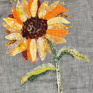 May include: A hand-stitched sunflower with yellow, orange, and brown fabric petals. The sunflower is on a gray linen background.