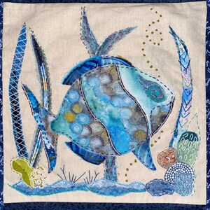 May include: A square fabric wall hanging featuring a central blue fish appliqué with intricate circular patterns and stitching. The scene includes tall blue seaweed, small gold bubbles, and textured coral elements on a light beige linen background.