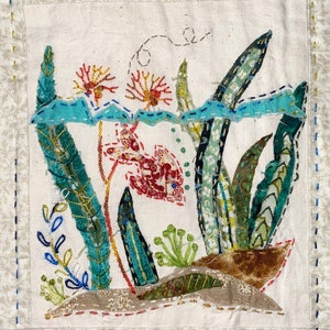 May include: A colorful embroidered artwork depicting a stylized fish swimming among green and blue plants. The artwork is stitched on a white fabric background.