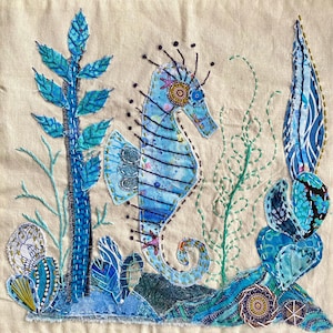 May include: A textile art piece on beige fabric featuring a central blue seahorse surrounded by embroidered aquatic plants and coral. The seahorse has intricate stitching and circular patterns, while the surrounding flora uses blue fabric scraps.