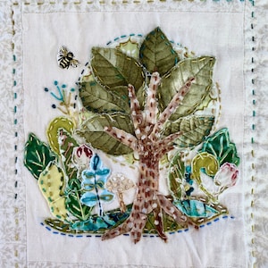 May include: A white fabric square with a stitched border features a colorful embroidered design of a tree with leaves, mushrooms, and a bee. The tree is brown with green leaves and a white trunk. The mushrooms are white with brown spots. The bee is yellow and black.