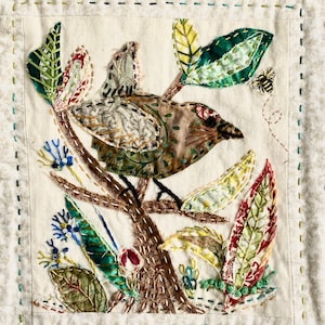 Puede incluir: Un pájaro marrón y verde bordado sobre un fondo de tela blanca con hojas verdes, amarillas y rojas. El pájaro está posado en una rama marrón con una abeja bordada en la esquina superior derecha.