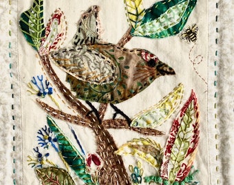May include: A brown and green bird embroidered on a white fabric background with green, yellow, and red leaves. The bird is perched on a brown branch with a bee embroidered on the upper right corner.