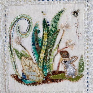 Puede incluir: Una tela blanca con hojas verdes bordadas, un conejo marrón y una abeja. La tela tiene un borde cosido.