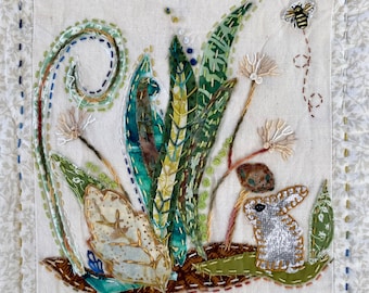 May include: A white fabric with embroidered green leaves, a brown rabbit, and a bee. The fabric has a stitched border.