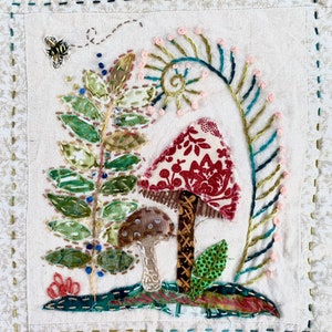 May include: A textile artwork depicting embroidered mushrooms, leaves, and a bee. The large mushroom has a red and white patterned cap. Green and blue embroidery details the leaves and stems. The background is cream with a floral pattern.