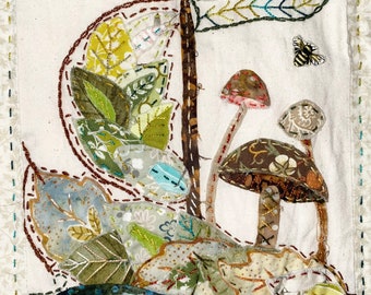 May include: A handmade fabric art piece depicting a mushroom with a brown cap and stem, surrounded by green and brown leaves. The piece is stitched with brown thread and has a white background.
