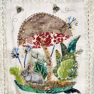 May include: A whimsical embroidered wall art featuring a mushroom with a red and white patterned cap, surrounded by green leaves and a small grey rabbit. The background is a cream-coloured fabric with a subtle texture.