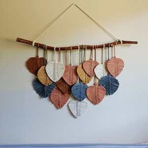 May include: A wall hanging made of macrame with a natural branch and a variety of colored heart shapes. The hearts are in shades of beige, brown, blue, and white.