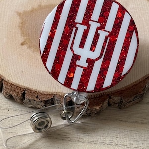 May include: A round badge reel with a red glitter background and white stripes. The badge reel features the white stylized letter "I" and "U" logo. The reel has a clear plastic strap and a metal clip.