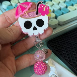 Peut inclure: Un enrouleur de badge en forme de crâne blanc rappelant un verre à cocktail tropical avec des paillettes roses, un parasol rose, une paille noire et une fleur d'hibiscus rose. Sous le crâne, deux perles rondes transparentes et rose vif.